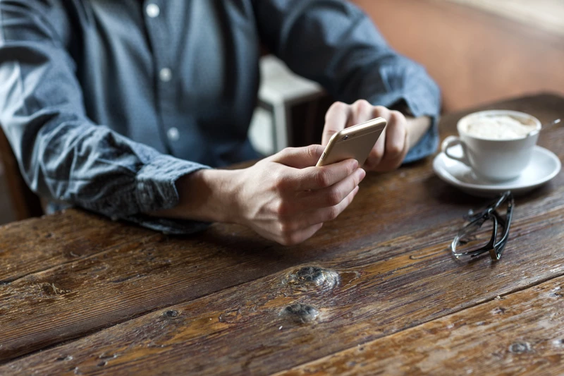 händer på en man som sitter vid ett bord med en kopp kaffe och som ska ringa ett samtal på sin mobil