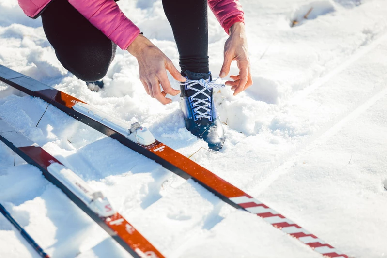 En person som tar på sig skidpjäxor bredvid ett par längdskidor i snön